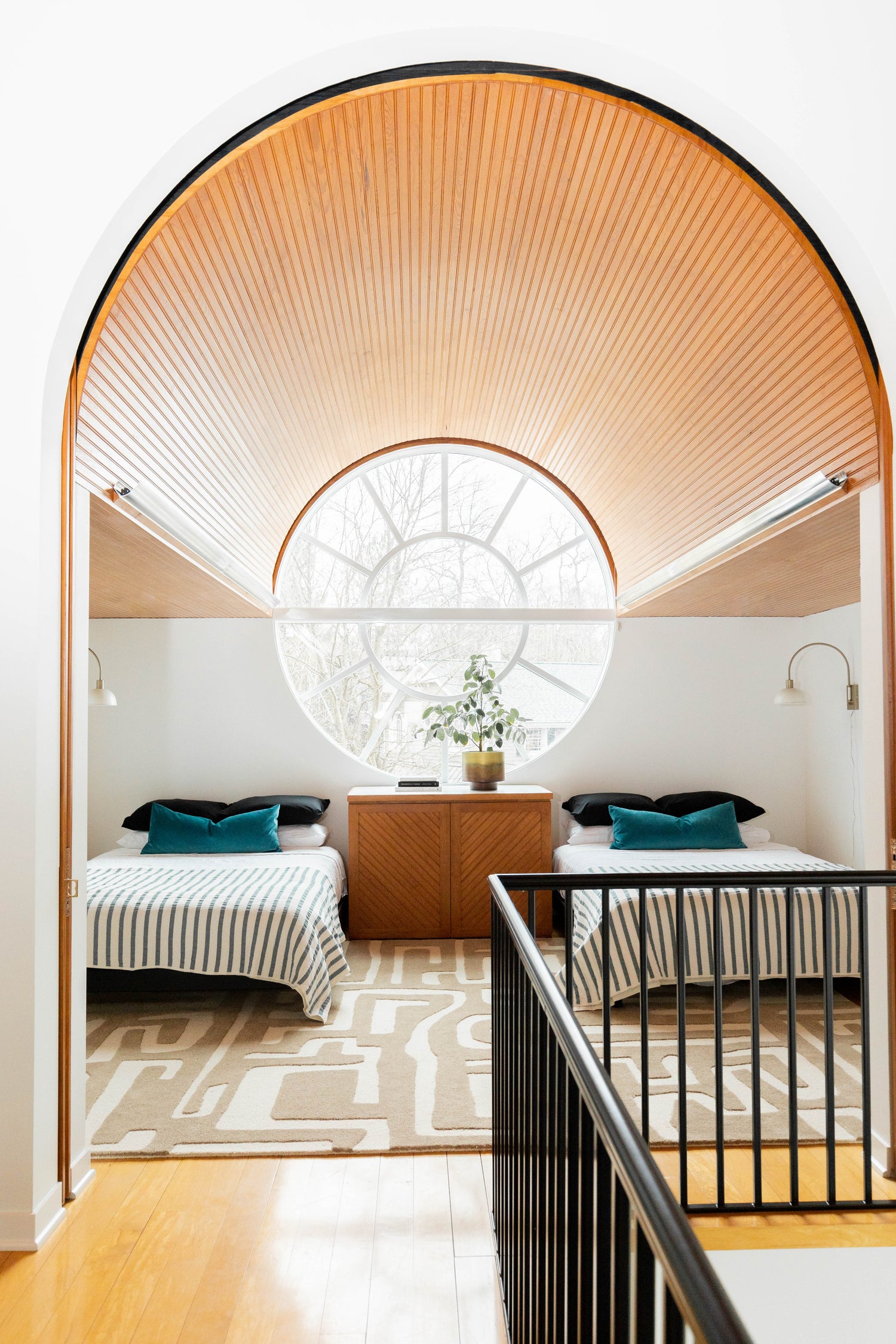 Arched wood ceiling, large round window grounded in the middle of the room, double queen bed set up with a pop of teal as a lumbar pillow, utilizing the walker noble rug from west elm.
