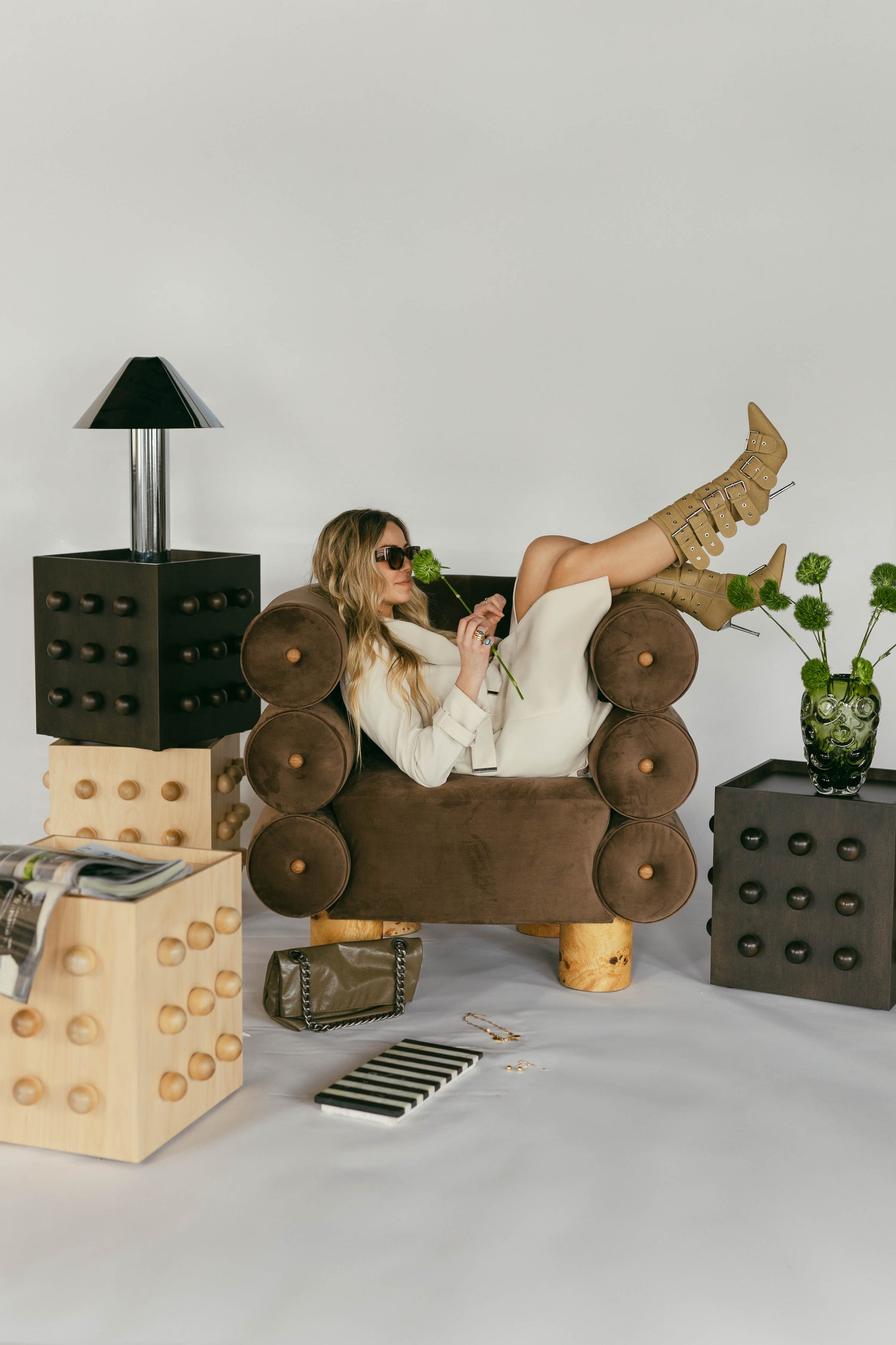 luxe furniture styled in a studio, brown velvet arm chair burl wood legs, dark and light accent maple tables, model styled in a trench coat with pastel ochre heeled buckle boots. accessories styled editorially around the pieces and floor.