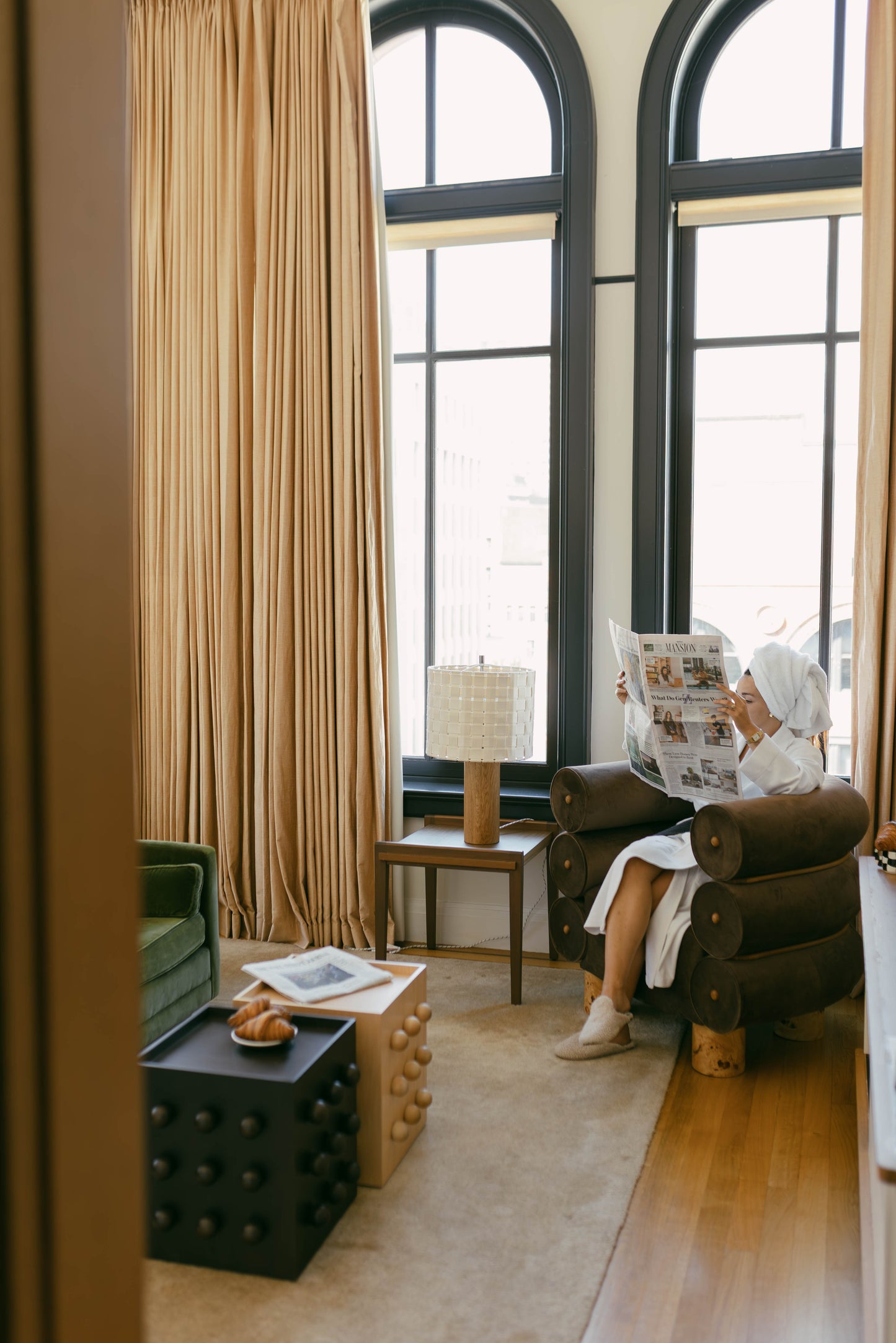 Living room with large windows, chocolate brown armchair, and person reading a newspaper with layered coffee table.