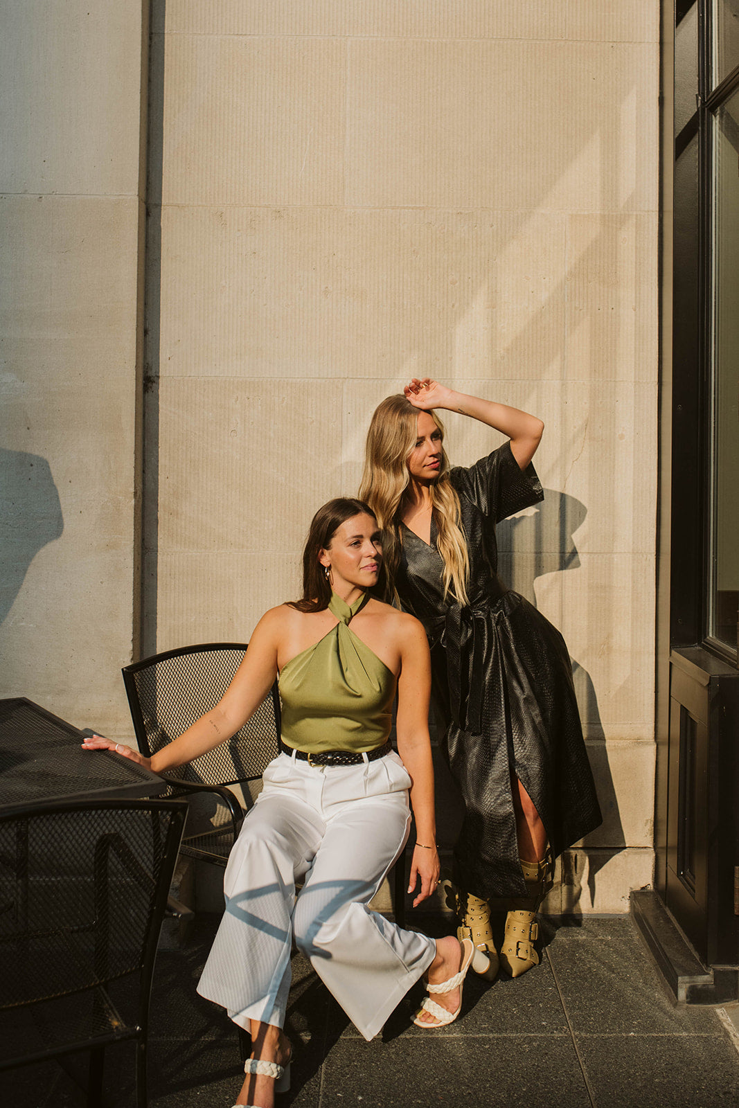 Luxury furniture designers styled for a brand shoot in the morning light against a concrete building sitting at an iron outdoor set. Styled in a black leather dress and pastel ochre buckle boots and a green neck tie top with white trousers and braided white heels.