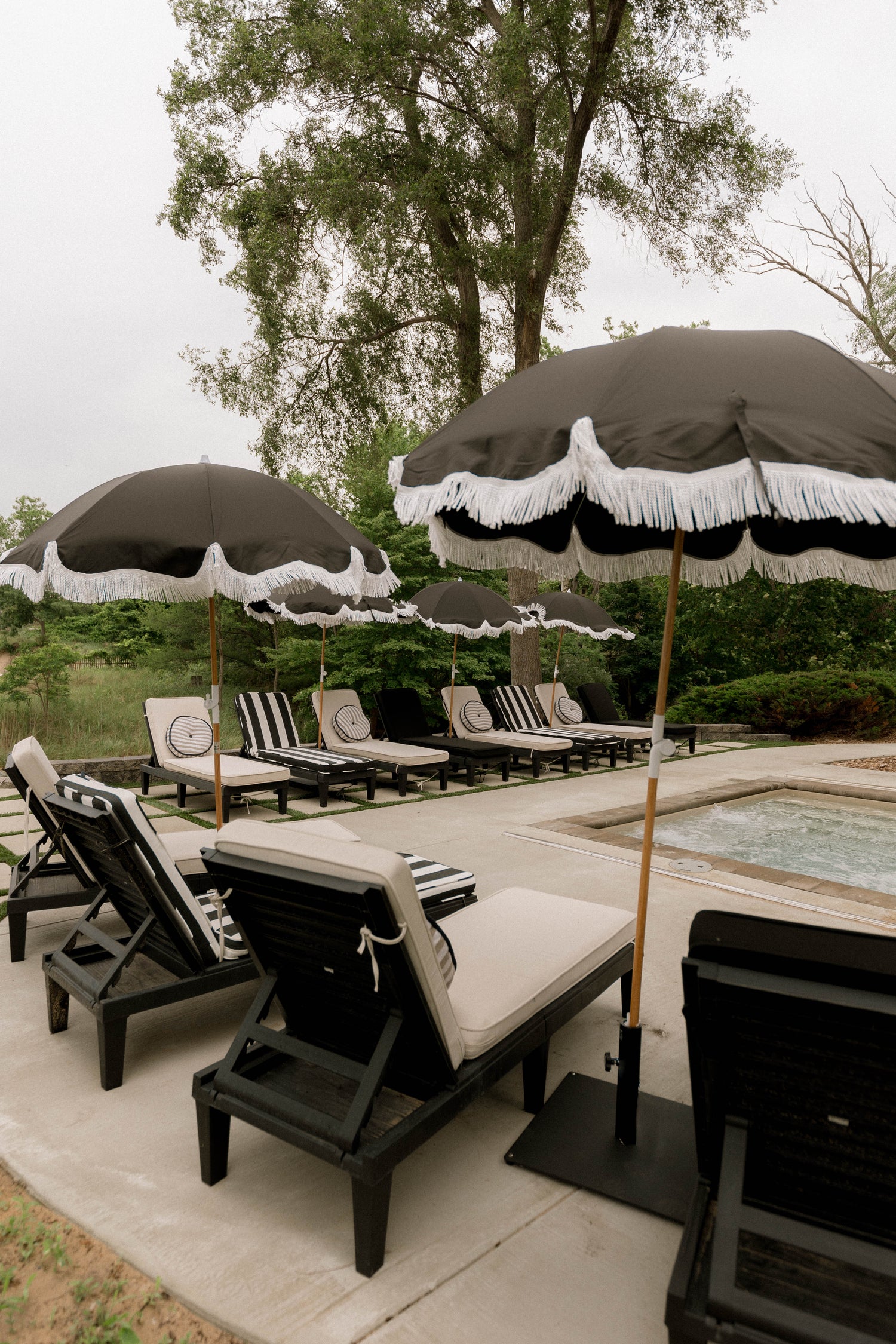 Pool side design around a inground spa with a mix of white, black and thick striped chaise cushions styled with striped round outdoor pillow, black luxury and pleasure umbrellas with white fringe.
