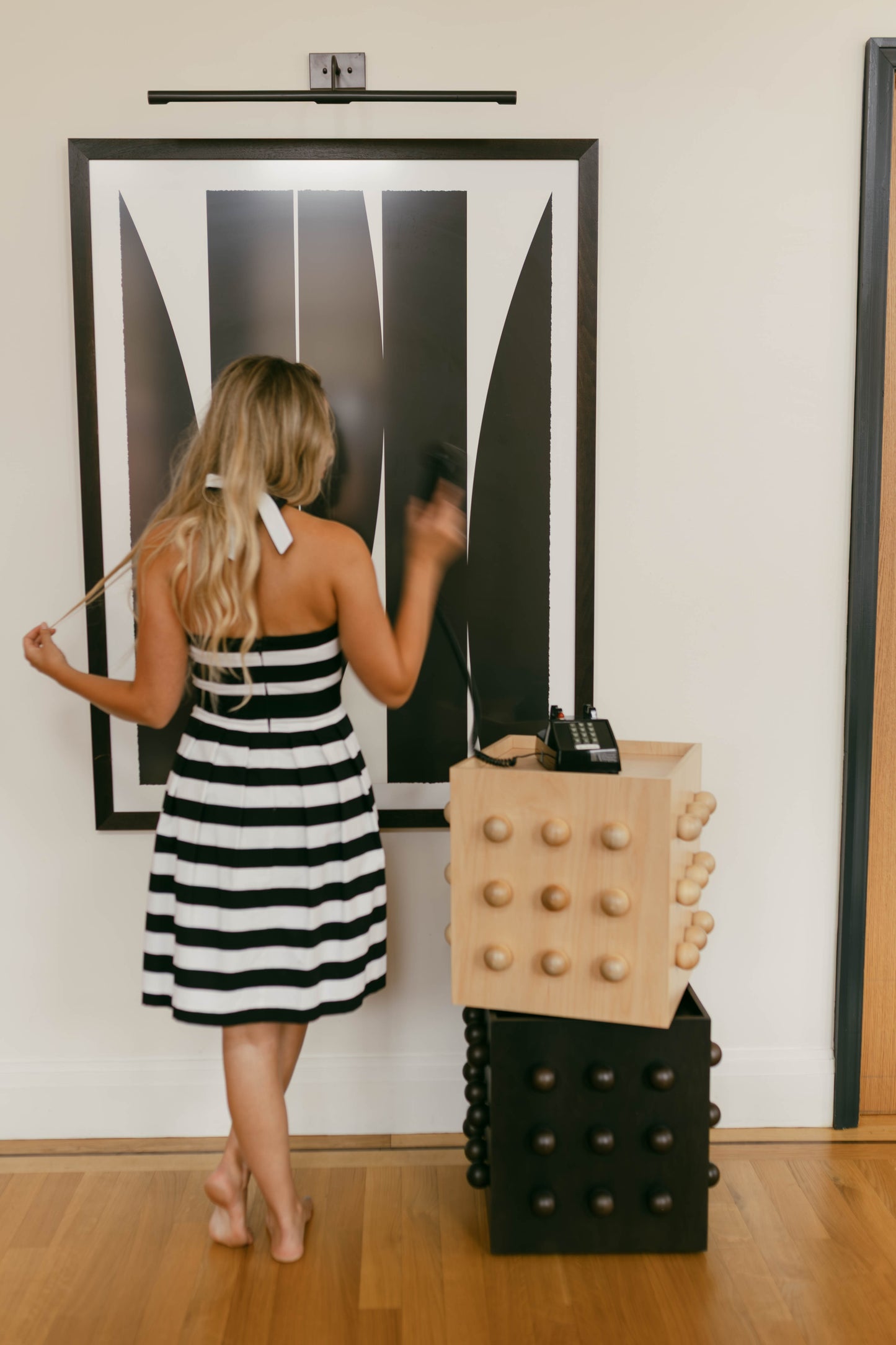 Woman in a black and white striped dress standing next to a geometric art piece on a wall.