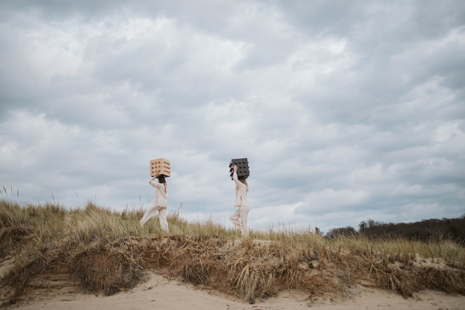 Two people carrying sculptural accent tables on beach under a cloudy sky