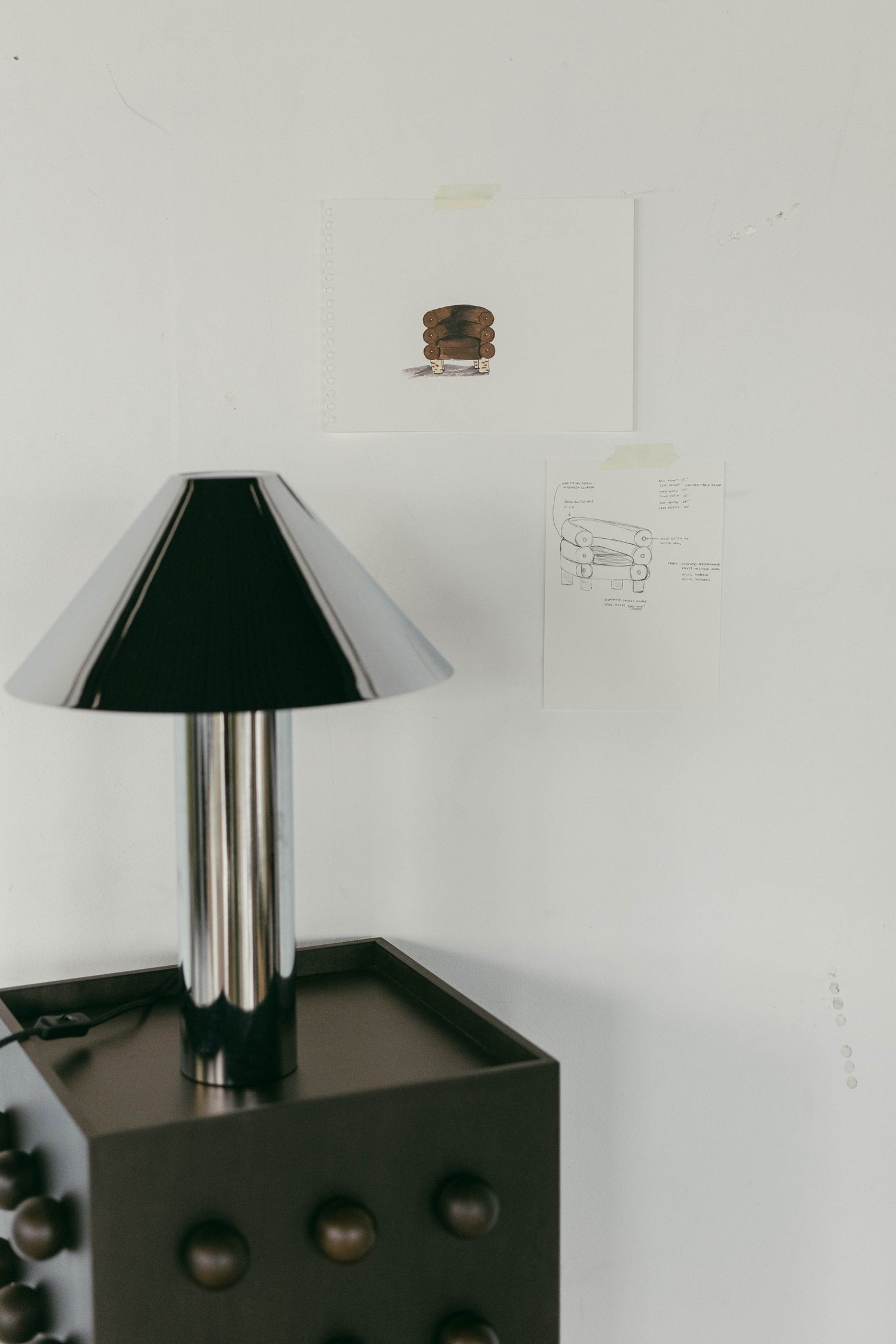 Concept drawings of lux velvet arm chair in chocolate taped to wall behind styled accent table with a chrome lamp. 